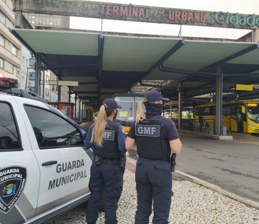 Duas guardas municipais uniformizadas e de costas ao lado de um carro da Guarda Municipal, elas estavam no terminal urbano de Florianópolis, reforçando a fiscalização das medidas do novo decreto.