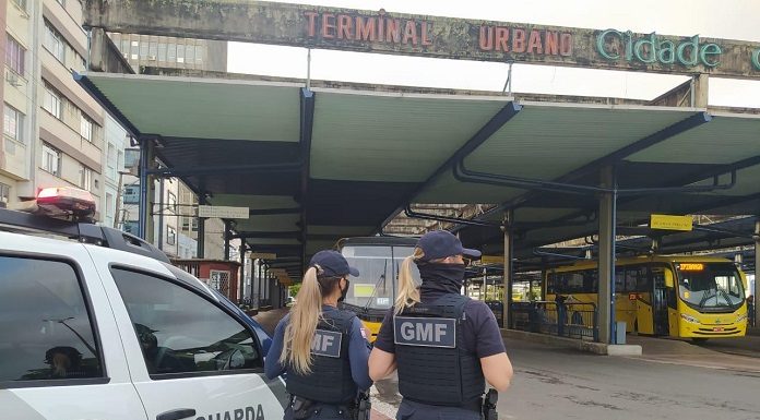 Duas guardas municipais uniformizadas e de costas ao lado de um carro da Guarda Municipal, elas estavam no terminal urbano de Florianópolis, reforçando a fiscalização das medidas do novo decreto.