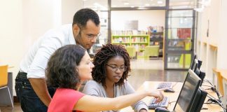 Uma mulher sentada aponta para a tela do computador enquanto outra mulher, sentada ao seu lado, digita no teclado. Atrás delas está um homem acompanhando o trabalho. A imagem ilustra o estágio de estudantes em empresas, como as de tecnologia, que abriram vagas em SC.