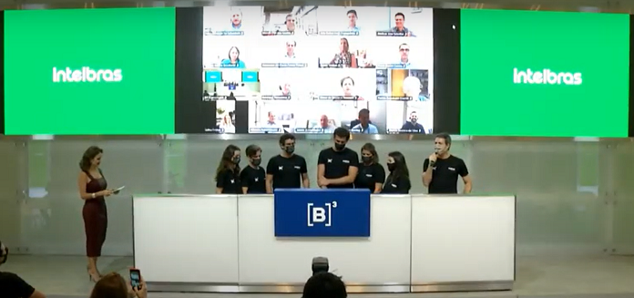 Seis representantes da Intelbras em evento que marca estreia da empresa na bolsa de valores. Ao fundo três telas, a do meio com participantes do evento em chamada de vídeo e as outras duas com cor verde e "Intelbras" escrito em branco.