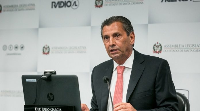 Deputado Julio Garcia tem pele branca e cabelo escuro penteado para trás, usa terno escuro com gravata vermelha, está a frente de microfones em cima de uma mesa e tem a sua frente também um computador e uma placa com seu nome, atrás um banner com logo da Alesc e rádio AL. Nesta semana o STF julgou a reclamação da Alesc procedente e Julio Garcia poderá retornar ao cargo.