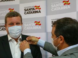 Moisés dá posse a novo secretário, Luciano Buligon, colocando um broche em sua lapela; painel de logos do governo sc ao fundo