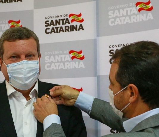 Moisés dá posse a novo secretário, Luciano Buligon, no Desenvolvimento Econômico Moisés dá posse a novo secretário, Luciano Buligon, colocando um broche em sua lapela; painel de logos do governo sc ao fundo