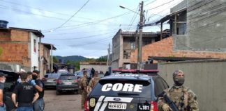 uma rua com um homem fardado e armado próximo a um carro da polícia civil, ao fundo um grupo de homens conversa e é possível ver outro grupo de homens fardados conversando. Nesta semana foi deflagrada a operação maserati.