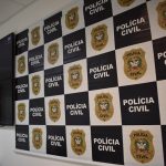 A imagem ilustra a prisão efetuado por policiais civis de um homem suspeito de estupro em São José. Painel com quadros brancos e brasão da polícia civil ao lado de quadros pretos com a escrita em branco de "polícia civil". Ao lado há um televisor.