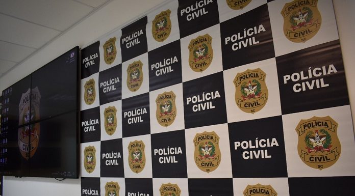 A imagem ilustra a prisão efetuado por policiais civis de um homem suspeito de estupro em São José. Painel com quadros brancos e brasão da polícia civil ao lado de quadros pretos com a escrita em branco de "polícia civil". Ao lado há um televisor.