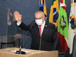 O prefeito de Itajaí de terno preto, gravata vermelha, óculos preto e máscara branca em frente a um microfone e com a mão erguida durante a posse. O MP pede a cassação do mandato do prefeito e vice por irregularidades nas eleições.