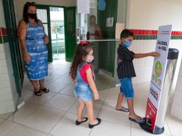 duas crianças usando máscaras em frente a totem de álcool em gel com a mãe em escola; período de pré-matrícula na rede estadual