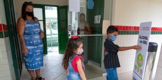 duas crianças usando máscaras em frente a totem de álcool em gel com a mãe em escola; período de pré-matrícula na rede estadual