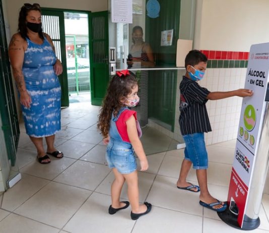 Pré-matrícula na rede estadual abre na internet na quarta e quinta-feira duas crianças usando máscaras em frente a totem de álcool em gel com a mãe em escola; período de pré-matrícula na rede estadual