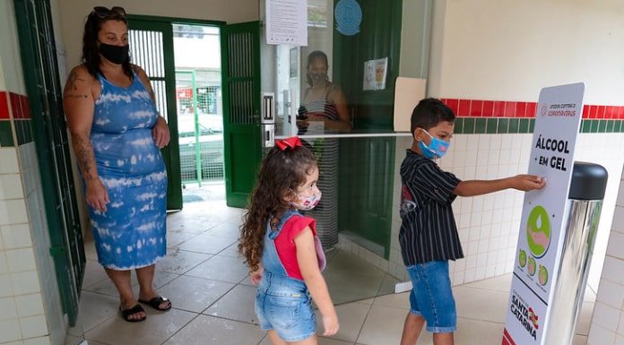 duas crianças usando máscaras em frente a totem de álcool em gel com a mãe em escola; período de pré-matrícula na rede estadual