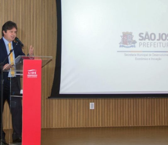 O secretário de desenvolvimento de São José, Marcelo Fett, à esquerda. À direita e mais ao fundo um telão em branco com o brasão de São José no centro. No evento, Fett apresentou o programa Avança São José de retomada econômica