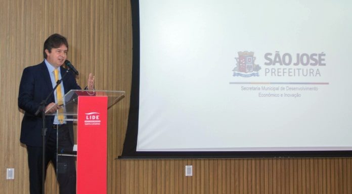 O secretário de desenvolvimento de São José, Marcelo Fett, à esquerda. À direita e mais ao fundo um telão em branco com o brasão de São José no centro. No evento, Fett apresentou o programa Avança São José de retomada econômica
