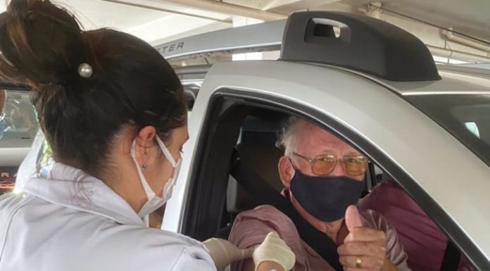 idosos sendo vacinado pela janela do carro por uma profissional de saúde; ele faz sinal de positivo para a foto