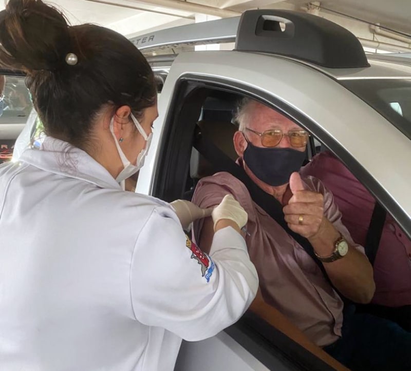 idosos sendo vacinado pela janela do carro por uma profissional de saúde; ele faz sinal de positivo para a foto