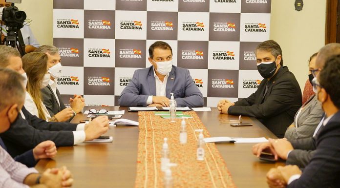 carlos moisés sentado na ponta de grande mesa; usa máscara; pessoas de máscara em volta; painel do governo ao fundo