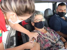 idosa recebe dose no braço por uma profissional de saúde; homem no banco do motorista; todos de máscara - biguaçu vacina idosos de 71 anos