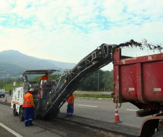 maquina recortando asfalto da via expressa sul e jogando detrito em caçamba de caminhão; operários ao lado; trânsito ao fundo