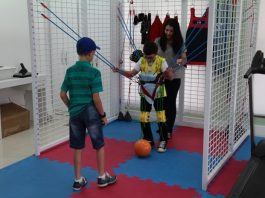 Paciente infantil do centro catarinense de reabilitação em atividade no local. Ele está com uma roupa amarelo e usa um colete com elásticos presos a grades laterais. Atrás do menino uma mulher adulta e na sua frente outra criança, há uma bola esportiva entre eles.