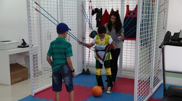 Paciente infantil do centro catarinense de reabilitação em atividade no local. Ele está com uma roupa amarelo e usa um colete com elásticos presos a grades laterais. Atrás do menino uma mulher adulta e na sua frente outra criança, há uma bola esportiva entre eles.