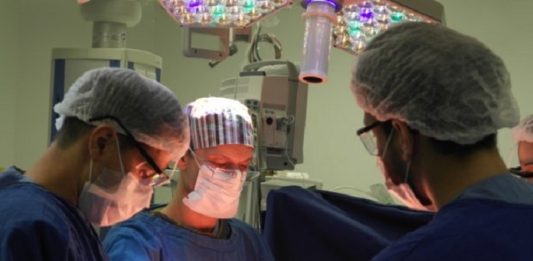 Três profissionais da saúde com roupas azuis, máscaras e proteção nos olhos e cabelos se concentram em um procedimento cirúrgico. As cirurgias eletivas em Santa Catarina estão suspensas na rede privada até o fim de março.