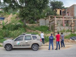Carro da PM estacionado em frente a uma área verde com construções irregulares, em São José. Quatro pessoas observam a área.