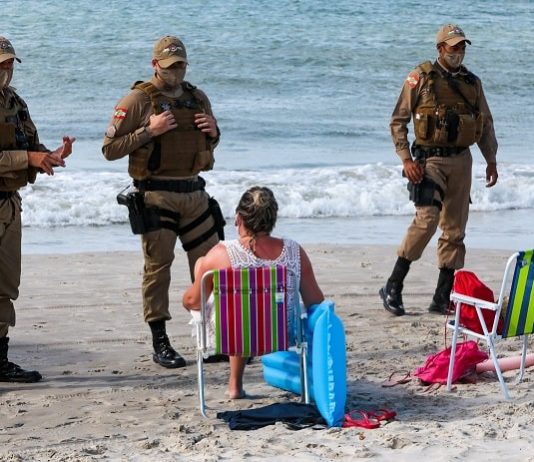 dois policiais usando máscara conversam com mulher sentada em cadeira de praia na beira do mar; outro policial de máscara ao lado - serviços que podem funcionar em sc no fim de semana