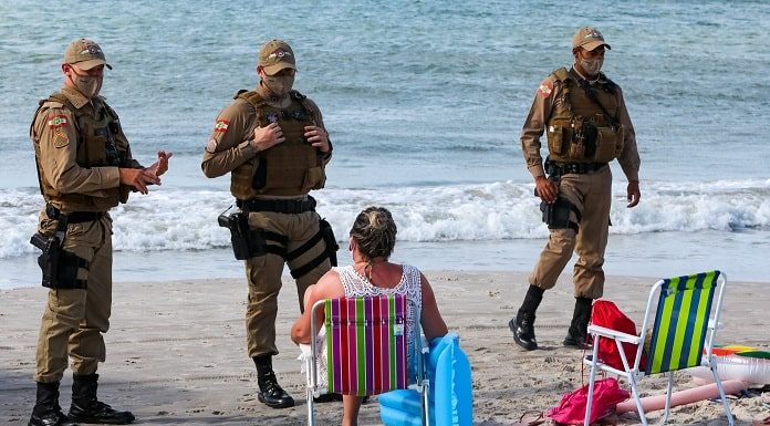 dois policiais usando máscara conversam com mulher sentada em cadeira de praia na beira do mar; outro policial de máscara ao lado - serviços que podem funcionar em sc no fim de semana