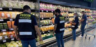 Quatro pessoas afastadas estão na frente da parte de frios de um supermercado, três delas usam colete de fiscalização do Procon. Eles também usam máscaras.