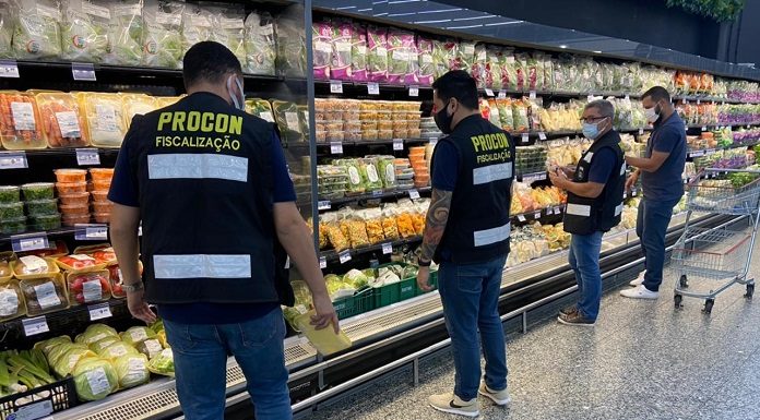 Quatro pessoas afastadas estão na frente da parte de frios de um supermercado, três delas usam colete de fiscalização do Procon. Eles também usam máscaras.