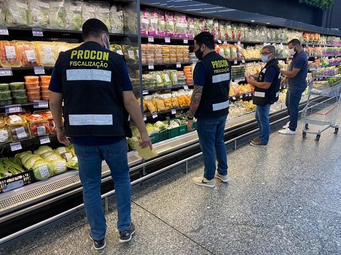 Quatro pessoas afastadas estão na frente da parte de frios de um supermercado, três delas usam colete de fiscalização do Procon. Eles também usam máscaras.