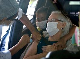 idosa sendo vacinada dentro de carro por profissional de saúde ao lado da porta aberta; pessoa abraça idosa por trás no carro