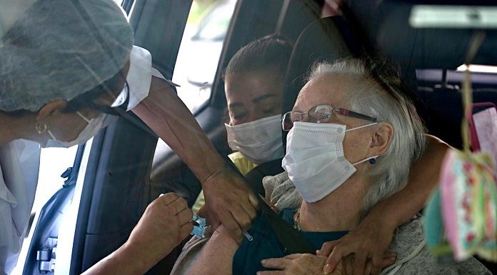 idosa sendo vacinada dentro de carro por profissional de saúde ao lado da porta aberta; pessoa abraça idosa por trás no carro