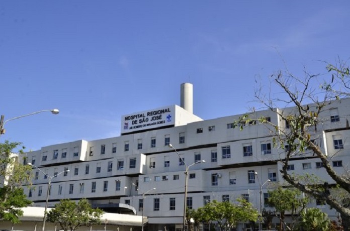 Visão lateral diagonal do Hospital Regional de São José, que registrou ontem o segundo apagão em março. A estrutura do hospital é grande, um prédio branco e em cima se lê a identificação. O céu está azul e há algumas árvores em volta.