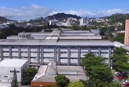 Profissionais do HU entram em greve por reajuste salarial Visão de cima do HU/UFSC. Aprecem algumas árvores e prédios ao fundo. O Hospital tem alguns prédios lado a lado, o que aparece na frente tem cor cinza e branco. Nesta semana, o HU fechou a emergência pediátrica para atender adultos em estado crítico da Covid.