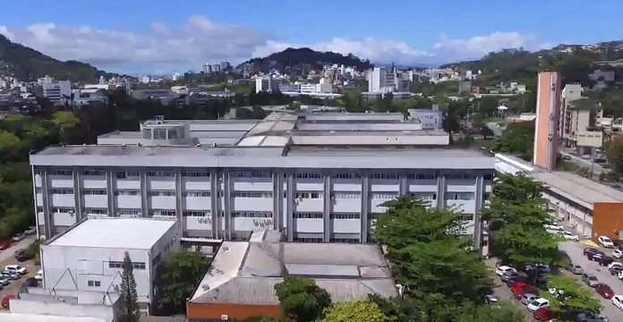 Visão de cima do HU/UFSC. Aprecem algumas árvores e prédios ao fundo. O Hospital tem alguns prédios lado a lado, o que aparece na frente tem cor cinza e branco. Nesta semana, o HU fechou a emergência pediátrica para atender adultos em estado crítico da Covid.