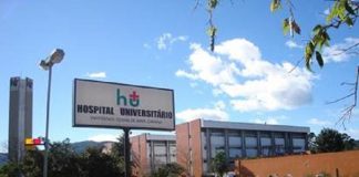 Visão do HU/UFSC onde aparece oe stacionamento com um uno branco parado, há uma placa que sinaliza que ali é o Hospital Universitário da UFSC, ao fundo se vê prédios alaranjados. Nesta semana, o HU fechou a emergência pediátrica para atender adultos em estado crítico da Covid.