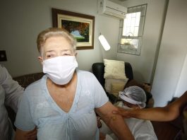 Senhora de cabelos loiros com máscara branca e blusa clara tem seu braço segurado por uma pessoa, além de estar acompanhada de mais duas pessoas de jaleco. Nesta semana a prefeitura de Florianópolis inicia a vacinação de idosos de 85 a 89 anos em casa.