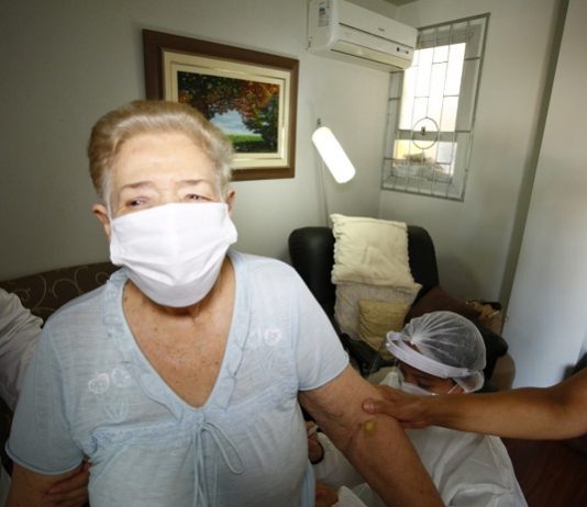 Idosos que não puderem ir aos drive-thrus serão vacinados em casa em Florianópolis Senhora de cabelos loiros com máscara branca e blusa clara tem seu braço segurado por uma pessoa, além de estar acompanhada de mais duas pessoas de jaleco. Nesta semana a prefeitura de Florianópolis inicia a vacinação de idosos de 85 a 89 anos em casa.