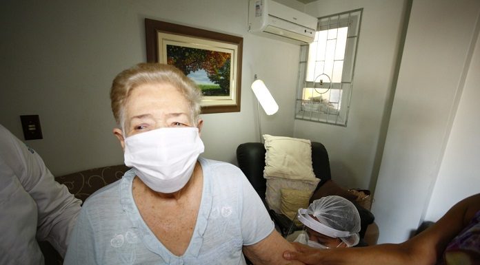 Senhora de cabelos loiros com máscara branca e blusa clara tem seu braço segurado por uma pessoa, além de estar acompanhada de mais duas pessoas de jaleco. Nesta semana a prefeitura de Florianópolis inicia a vacinação de idosos de 85 a 89 anos em casa.