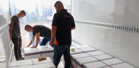 três homens andam em meios às caixas de isopor onde estão as doses de vacina dentro de um frigorífico