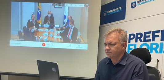gean loureiro sentado em ponta de mesa com notebook à frente e painel com reunião virtual ao fundo