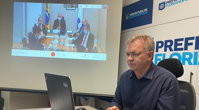 gean loureiro sentado em ponta de mesa com notebook à frente e painel com reunião virtual ao fundo