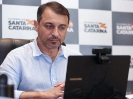 carlos moisés olha para um notebook; ao fundo painel de logos do governo