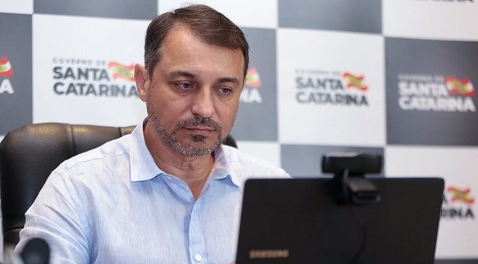 carlos moisés olha para um notebook; ao fundo painel de logos do governo