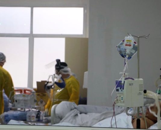 dois profissionais de saúde vistos de costas dentro de hospital; eles usam epi. em primeiro plano paciente intubado - sc tem fila de espera de uti de quase 400 pacientes