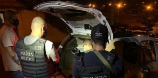 Duas pessoas de costas usam coletes que identificam a Polícia Civil. Na frente deles o porta malas aberto de um carro, dentro está o suspeito do homicídio contra o comerciante de Itapema.