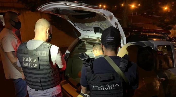 Duas pessoas de costas usam coletes que identificam a Polícia Civil. Na frente deles o porta malas aberto de um carro, dentro está o suspeito do homicídio contra o comerciante de Itapema.