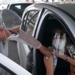 profissional de saúde segura algodão no braço de senhora dentro de carro; ela usa mácara e face shield; o homem usa diversos EPI