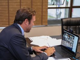 O presidente da OAB/SC de terno escuro em frente ao computador em reunião virtual. Sob a mesa papeis e ao fundo uma janela parte fechada com cortina e parte aberta.